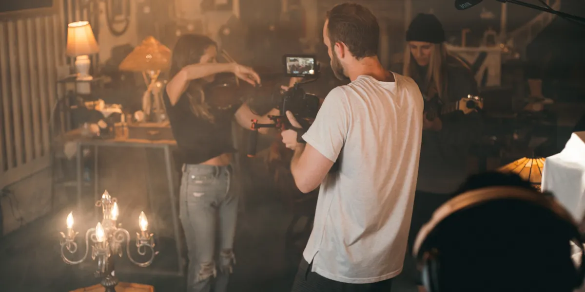 A camera operator films a violinist in a warm, lamp-lit room while two other crew members work behind him